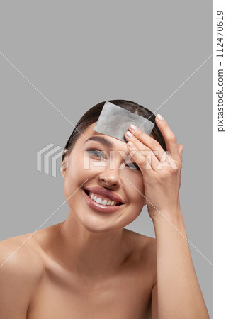 Face skin care. Smiling woman using facial oil blotting paper portrait. Closeup of beautiful happy girl model with natural makeup using oil absorbing sheets, beauty product 112470619