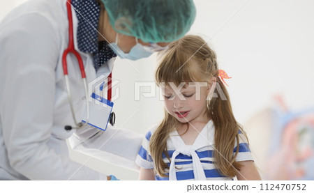 Doctor pediatrician in protective medical mask talking to little girl patient in clinic 112470752