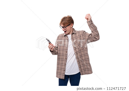 young handsome red-haired guy in a plaid shirt joyfully looks at the phone young handsome red-haired guy in a plaid shirt joyfully looks at the phone 112471727