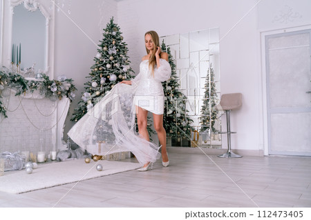 The blonde in the Christmas room. A beautiful blonde woman in a shiny light short dress with a train stands in a beautiful bright room decorated with a festive interior with a Christmas tree. The blonde in the Christmas room. A beautiful blonde woman in a shiny light short dress with a train stands in a beautiful bright room decorated with a festive interior with a Christmas tree. 112473405