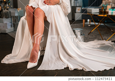 Pregnant Women legs christmas tree. Cropped photo of beautiful elegant female legs in white high-heeled shoes and a white dress, a woman sits against the background of a Christmas tree with gifts. Pregnant Women legs christmas tree. Cropped photo of beautiful elegant female legs in white high-heeled shoes and a white dress, a woman sits against the background of a Christmas tree with gifts. 112473411