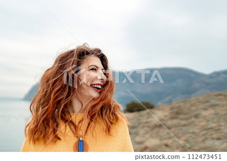 Portrait windswept hair happy woman against a backdrop of mountains and sea. Daylight illuminates the tranquil outdoor setting 112473451
