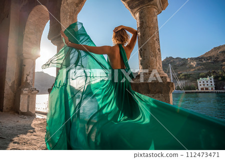 Woman dress sea columns. Rear view of a happy blonde woman in a long mint dress posing against the backdrop of the sea in an old building with columns. 112473471