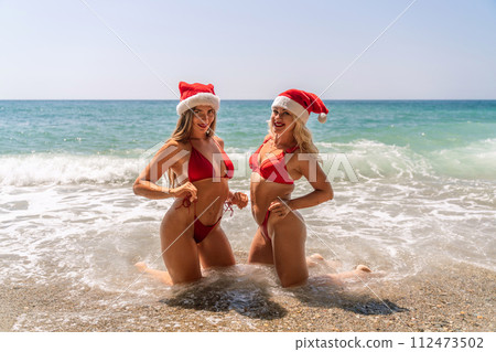 Women Santa hats ocean play. Seaside, beach daytime, enjoying beach fun. Two women in red swimsuits and Santa hats are enjoying themselves in the ocean waves. Women Santa hats ocean play. Seaside, beach daytime, enjoying beach fun. Two women in red swimsuits and Santa hats are enjoying themselves in the ocean waves. 112473502