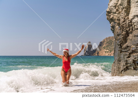 A woman in Santa hat on the seashore, dressed in a red swimsuit. New Year's celebration in a hot country. A woman in Santa hat on the seashore, dressed in a red swimsuit. New Year's celebration in a hot country. 112473504