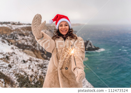Outdoor winter portrait of happy smiling woman, light faux fur coat holding heart sparkler, posing against sea and snow background 112473524