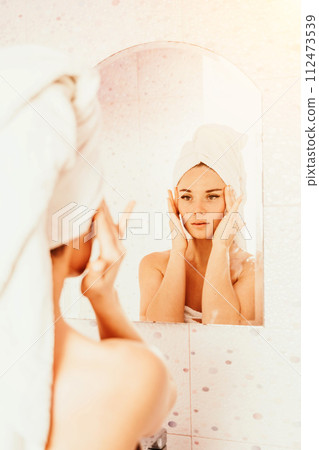 Young beautiful woman using face cream moisturizing lotion after bath. Pretty attractive girl in a towel on her head stands in front of a mirror in a home bathroom. Daily hygiene and skin care Young beautiful woman using face cream moisturizing lotion after bath. Pretty attractive girl in a towel on her head stands in front of a mirror in a home bathroom. Daily hygiene and skin care 112473539