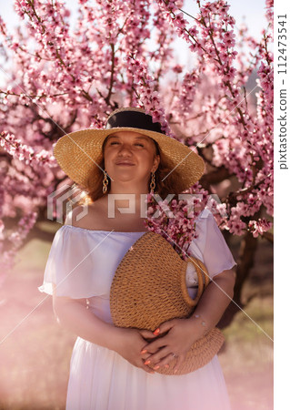 Woman blooming peach orchard. Against the backdrop of a picturesque peach orchard, a woman in a long white dress and hat enjoys a peaceful walk in the park, surrounded by the beauty of nature. Woman blooming peach orchard. Against the backdrop of a picturesque peach orchard, a woman in a long white dress and hat enjoys a peaceful walk in the park, surrounded by the beauty of nature. 112473541