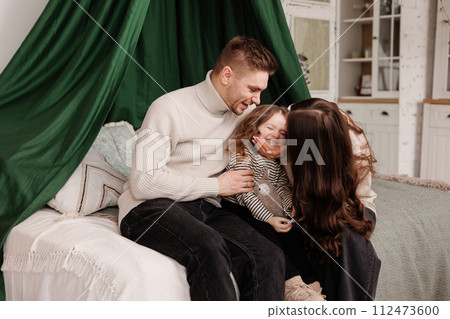 Happy family day. Young loving parents, mom dad with cute preschool little child daughter having fun, embracing at cozy bedroom on bed looking at camera. Parenthood childhood concept. 112473600