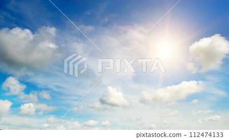 There are white cumulus clouds and a bright sun in the blue sky. 112474133