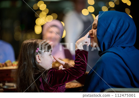In a heartwarming scene, a happy European Islamic family and a young girl delightfully engage with her mother while eagerly anticipating their iftar meal, radiating joy, love, and familial bonding 112475001