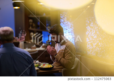 In a modern restaurant setting, a European Islamic family comes together for iftar during Ramadan, engaging in prayer before the meal, uniting tradition and contemporary practices in a celebration of In a modern restaurant setting, a European Islamic family comes together for iftar during Ramadan, engaging in prayer before the meal, uniting tradition and contemporary practices in a celebration of 112475063