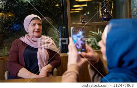 In a modern restaurant during the holy month of Ramadan, a woman in a hijab captures a moment with her mother using a smartphone, epitomizing the blending of tradition and technology in familial bonds In a modern restaurant during the holy month of Ramadan, a woman in a hijab captures a moment with her mother using a smartphone, epitomizing the blending of tradition and technology in familial bonds 112475068