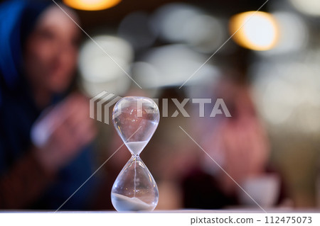 A symbolic hourglass counts down the time to iftar meal, while in the background, an Islamic family devoutly prays, capturing a poignant moment of spiritual connection and anticipation during Ramadan A symbolic hourglass counts down the time to iftar meal, while in the background, an Islamic family devoutly prays, capturing a poignant moment of spiritual connection and anticipation during Ramadan 112475073