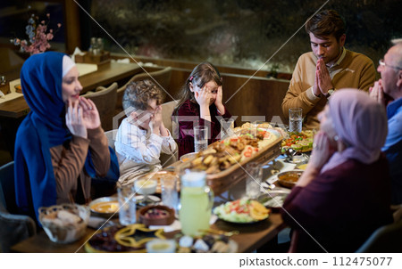 In a modern restaurant setting, a European Islamic family comes together for iftar during Ramadan, engaging in prayer before the meal, uniting tradition and contemporary practices in a celebration of 112475077