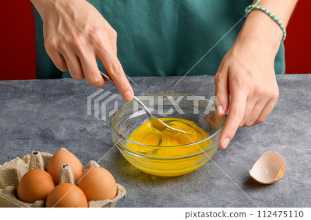 Female hands beat eggs with fork in a glass bowl on a gray kitchen table 112475110