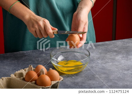 A woman's hand cracking an egg with knife into a clear glass bowl in the gray kitchen table A woman's hand cracking an egg with knife into a clear glass bowl in the gray kitchen table 112475124