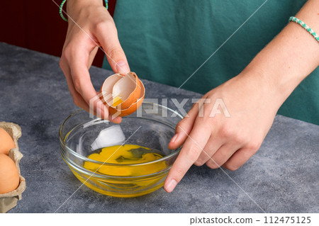 A woman's hand holding the eggshells of breked egg into a clear glass bowl in the gray kitchen table 112475125