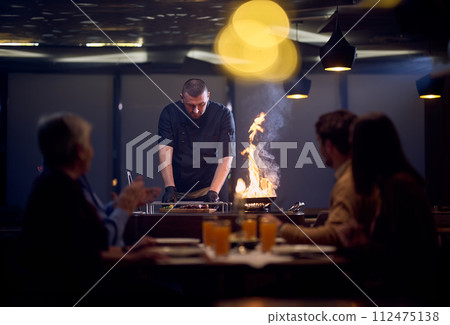 In a restaurant setting, a professional chef presents a sizzling steak cooked over an open flame, while an European Muslim family eagerly awaits their iftar meal during the holy month of Ramadan In a restaurant setting, a professional chef presents a sizzling steak cooked over an open flame, while an European Muslim family eagerly awaits their iftar meal during the holy month of Ramadan 112475138