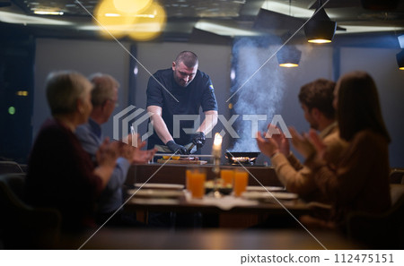 In a restaurant setting, a professional chef presents a sizzling steak cooked over an open flame, while an European Muslim family eagerly awaits their iftar meal during the holy month of Ramadan 112475151