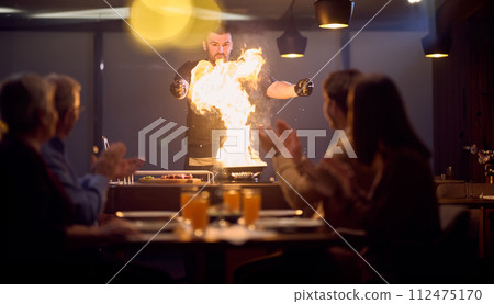 In a restaurant setting, a professional chef presents a sizzling steak cooked over an open flame, while an European Muslim family eagerly awaits their iftar meal during the holy month of Ramadan In a restaurant setting, a professional chef presents a sizzling steak cooked over an open flame, while an European Muslim family eagerly awaits their iftar meal during the holy month of Ramadan 112475170