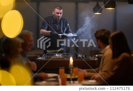 In a restaurant setting, a professional chef presents a sizzling steak cooked over an open flame, while an European Muslim family eagerly awaits their iftar meal during the holy month of Ramadan 112475178