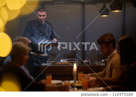 In a restaurant setting, a professional chef presents a sizzling steak cooked over an open flame, while an European Muslim family eagerly awaits their iftar meal during the holy month of Ramadan In a restaurant setting, a professional chef presents a sizzling steak cooked over an open flame, while an European Muslim family eagerly awaits their iftar meal during the holy month of Ramadan 112475189