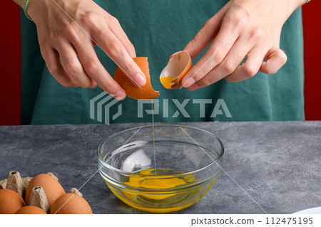 A woman's hand holding the eggshells of breked egg into a clear glass bowl in the gray kitchen table 112475195