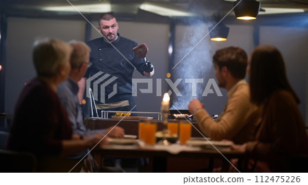 In a restaurant setting, a professional chef presents a sizzling steak cooked over an open flame, while an European Muslim family eagerly awaits their iftar meal during the holy month of Ramadan In a restaurant setting, a professional chef presents a sizzling steak cooked over an open flame, while an European Muslim family eagerly awaits their iftar meal during the holy month of Ramadan 112475226