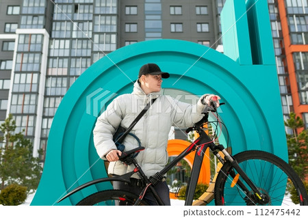 A young Caucasian man with a gray jacket and black cap spends time outdoors with a bicycle A young Caucasian man with a gray jacket and black cap spends time outdoors with a bicycle 112475442