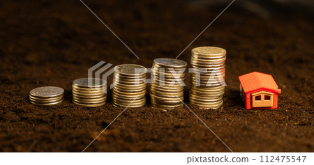 Money lies in piles on the ground next to a small house, symbolizing a loan or mortgage to purchase a house with land. Buying a house. There are coins on the black soil, and a house stands nearby. Money lies in piles on the ground next to a small house, symbolizing a loan or mortgage to purchase a house with land. Buying a house. There are coins on the black soil, and a house stands nearby. 112475547
