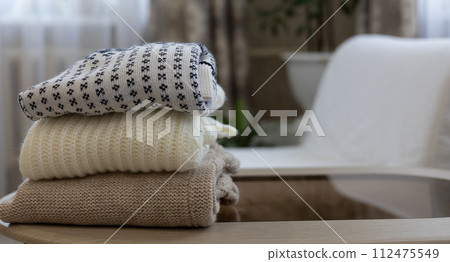 A stack of knitted clothes is folded on a table in the living room. Dismantling the closet from old, boring clothes. Second life for old clothes, recycling, charity, donation. A stack of knitted clothes is folded on a table in the living room. Dismantling the closet from old, boring clothes. Second life for old clothes, recycling, charity, donation. 112475549