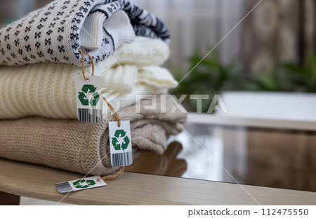 Clothes lie in a neat stack with a recycling sign on the table. Copy space, place for inscription. A symbol of conscious use of things. A second life for old clothes. Recycling, sale of used clothes. Clothes lie in a neat stack with a recycling sign on the table. Copy space, place for inscription. A symbol of conscious use of things. A second life for old clothes. Recycling, sale of used clothes. 112475550
