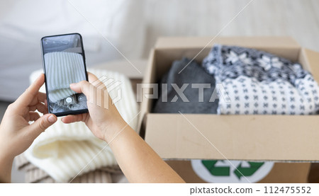 Having sorted out her clothes, the woman photographs them for sale and puts them in a box. Close-up of hands with a phone taking photos of a stack of clothes in the living room. Having sorted out her clothes, the woman photographs them for sale and puts them in a box. Close-up of hands with a phone taking photos of a stack of clothes in the living room. 112475552