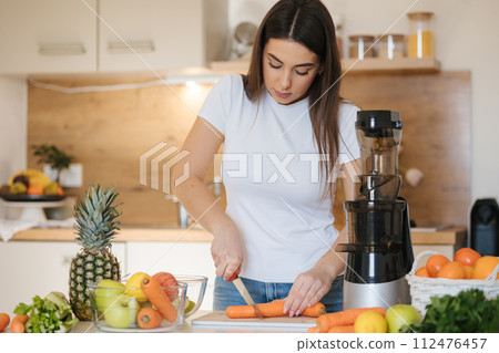 Attractive young woman cutting carrot in kitchen. Female preparing vegetables and fruits for juice. Home made fresh. Juicer Attractive young woman cutting carrot in kitchen. Female preparing vegetables and fruits for juice. Home made fresh. Juicer 112476457