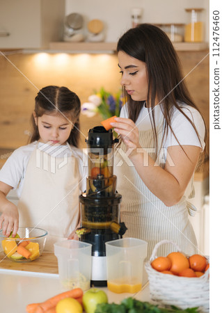 Adorable five year old girl putting with her mom fruits into juicer. Two females into aprons. 112476460