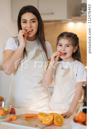 Young mom and cute daughter in aprons try carrots in the kitchen. Happy family sliced fruits and vegetables at home Young mom and cute daughter in aprons try carrots in the kitchen. Happy family sliced fruits and vegetables at home 112476463