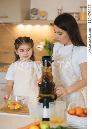 Adorable five year old girl putting with her mom fruits into juicer. Two females into aprons. Adorable five year old girl putting with her mom fruits into juicer. Two females into aprons. 112476465