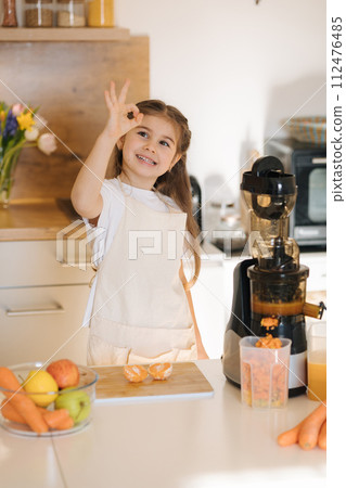 Portrait of cute five year old girl in apron preparing organic fruits and vegetables for fresh juice. Homemade vegan beverage from juicer 112476485