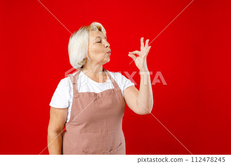 Elderly chef cooker housekeeper housewife woman in apron show sign of good taste, Bellissimo gesture, kiss fingers isolated plain on red color background. 112478245