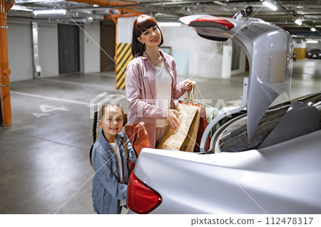 Mother and little daughter putting colorful bags with clothes into trunk of car. 112478317