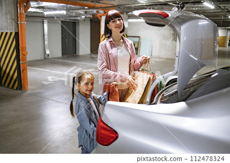 Mother and little daughter putting colorful bags with clothes into trunk of car. 112478324