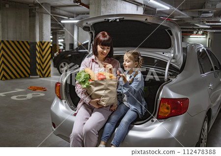 Mother and daughter sitting in trunk of vehicle holding paper bag with groceries 112478388