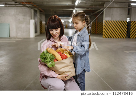 Mother with preschool daughter holding paper bag with vegetables in hands 112478416