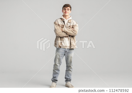 A young man in a tan jacket and blue jeans stands with his arms crossed 112478592