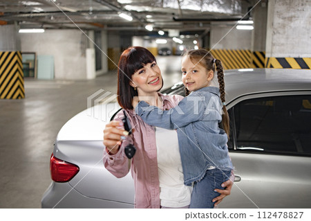 Happy Caucasian woman holding female kid and car keys in underground parking lot Happy Caucasian woman holding female kid and car keys in underground parking lot 112478827