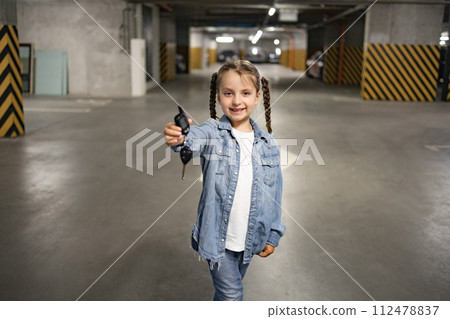 Little girl standing in underground parking lot in casual clothes holding keys 112478837