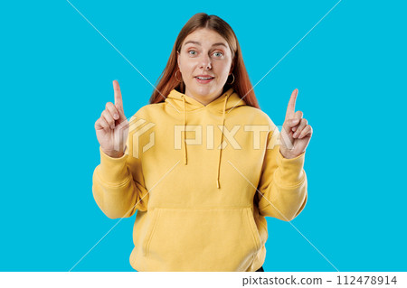 Cheerful happy female model pointing fingers up to shop promotion banner, smiling camera, studio portrait. 30s Woman pointing fingers at copy space isolated over blue background, surprised face. Cheerful happy female model pointing fingers up to shop promotion banner, smiling camera, studio portrait. 30s Woman pointing fingers at copy space isolated over blue background, surprised face. 112478914