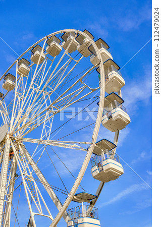 Bright white Ferris wheel on sunny day and clear blue sky with clouds, Cascais, Portugal. People enjoying rides on holiday, vertical shot, joy and leisure in sunshine. Bright white Ferris wheel on sunny day and clear blue sky with clouds, Cascais, Portugal. People enjoying rides on holiday, vertical shot, joy and leisure in sunshine. 112479024