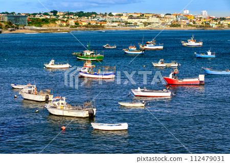 Colorful view of small fishing boats on sunny day in bay of Cascais, Portugal, quiet and luxurious seaside town near Lisbon with beaches and small port. Clear blue sky, clouds, sunshine, calm ocean 112479031